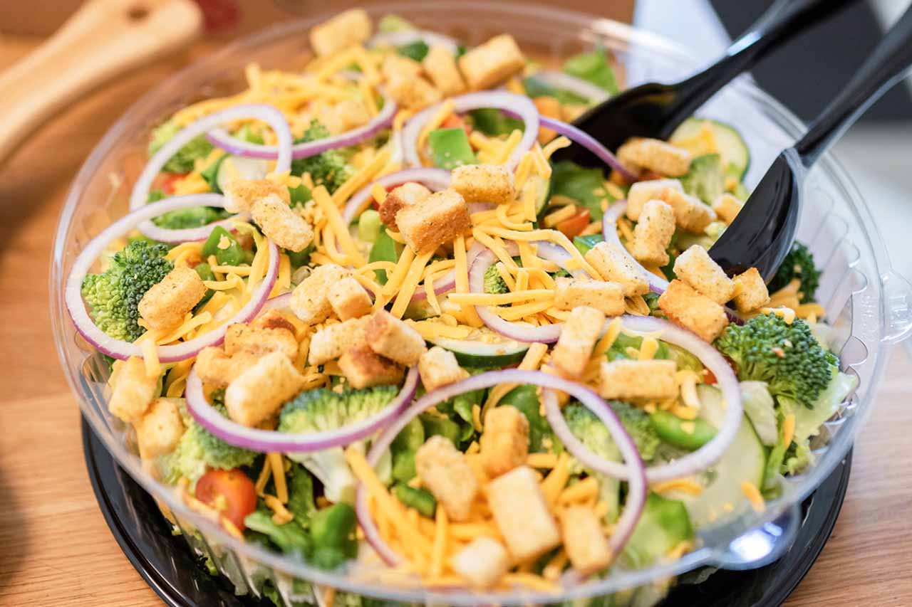 Small group garden salad with broccoli, croutons, cucumbers, green peppers, red onions, shredded cheddar cheese shown with tongs.